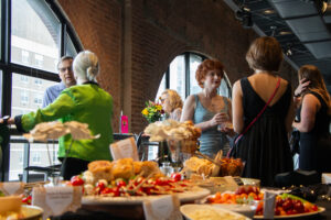 A table of food. In the background, people mingle.