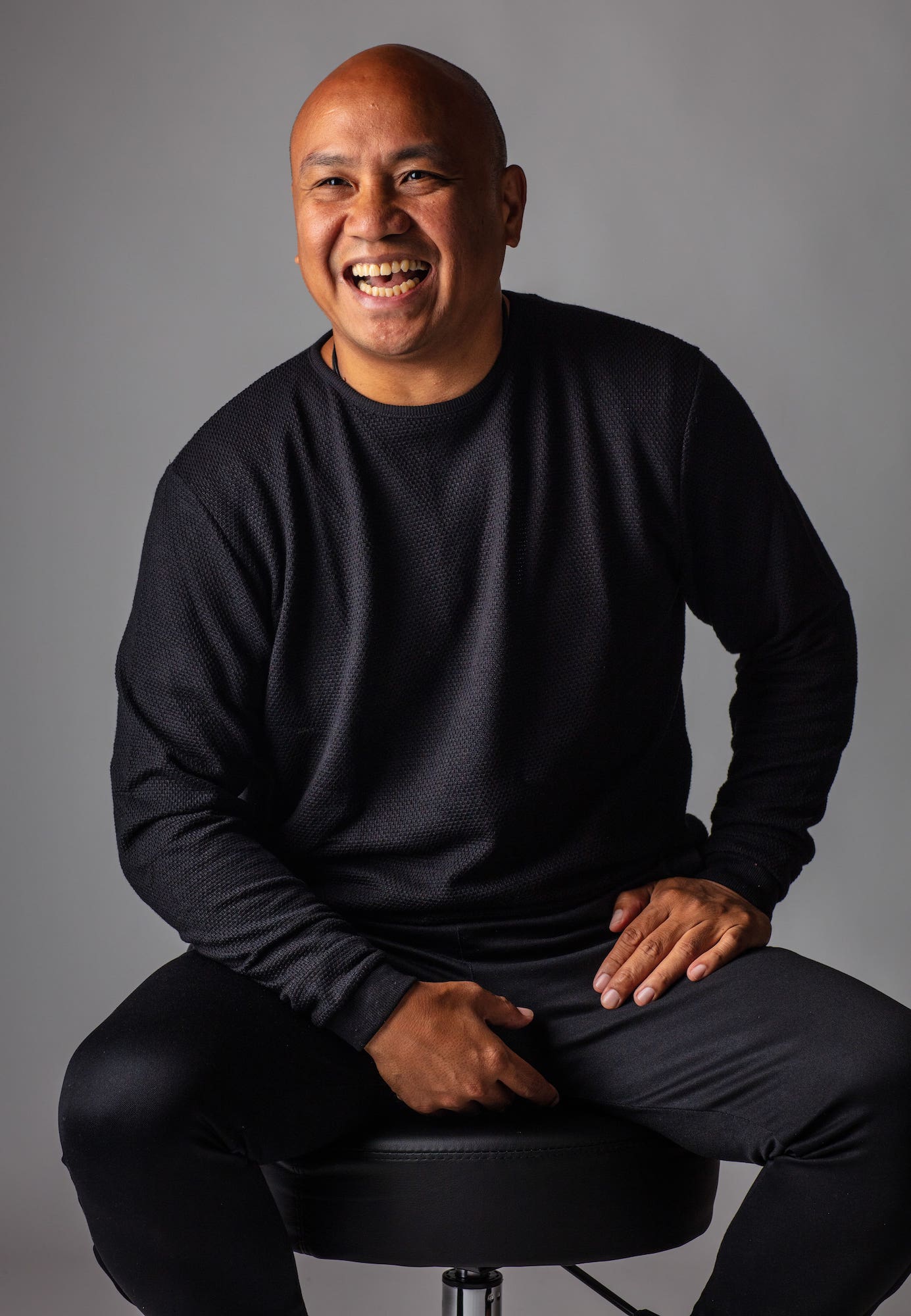 Photographic portrait of Patrick Rosal, a middle-aged Filipno American man. Rosal wears a black crewneck sweater and black pants. He is sitting on a stool, looking to his left, and is captured mid-laugh.