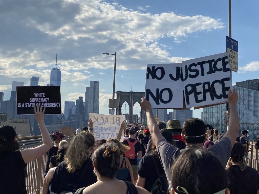 Image of a protest on the Brooklyn Bridge in NYC, with someone holding up a sign saying "No Justice, No Peace."