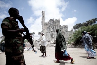 Mogadishu: a man holds an assault rifle while people walk by, with a crumbling building in the background