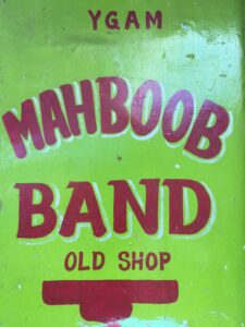 Image of an orange-green and red sign, reading "Ygam Mahboob Band Old Shop."