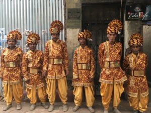 Image of 6 members of a marching band from India dressed in orange and yellow posing for a photo.