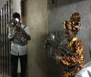 Image of two men playing brass instruments, one wearing a white shirt, the other dressed in yellow and orange.