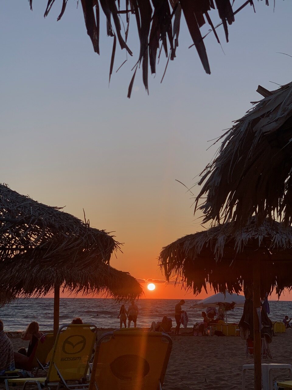 Image of the sun setting over the ocean with umbrellas made of twine, chairs, and people.