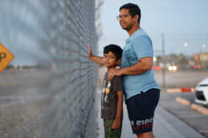 A man and a boy face a metal fence, with the boy looking at us.