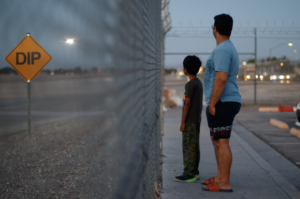 A man and a boy stand by a fence and look away from us.