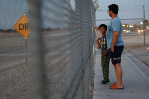 A boy and a man stand in front of a metal fence, looking at one another.