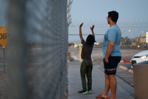 A man and a boy stand in front of a metal fence. The boy holds his hands to the sky. They look away.