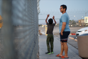 A boy and a man stand in front of a metal fence with a boy holding his hands to the sky.