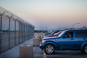 Woman sitting in a blue car parked in front of a metal fence.