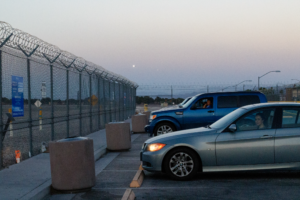 Woman sitting in a blue car parked in front of a metal fence next to another woman sitting in a grey car.