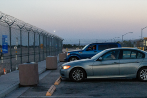 Woman sitting in a blue car parked in front of a metal fence next to another woman sitting in a grey car.