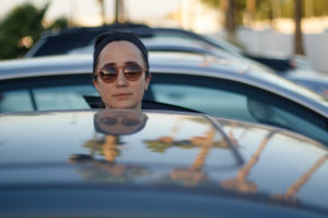 Image of a woman sticking her head from behind a car with palm trees in the background.