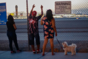 Three people and a dog facing a metal fence with an airport on the other side.