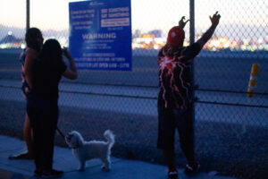 Three people and a dog facing a metal fence with an airport on the other side.