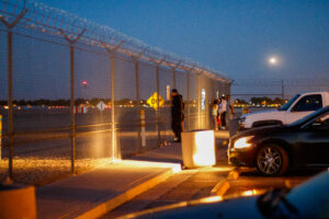 A person stands in front of a metal fence in the twilight.