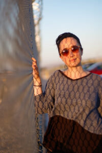Woman stands next to a chainlink fence in the sunlight.