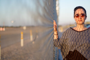 Image of a woman standing next to a chainlink fence in the sunlight.