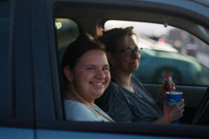 A woman and a girl sit in a dark blue car in a parking lot.