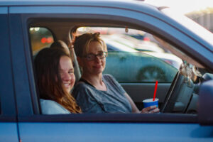 A woman and a girl sit in a dark blue car in a parking lot.