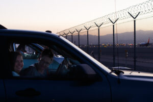 A woman and a girl sit in a dark blue car in a parking lot.