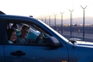 A woman and a girl sit in a dark blue car in a parking lot.