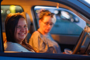 A woman and a girl sit in a dark blue car in a parking lot.