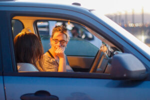 A woman and a girl sit in a dark blue car in a parking lot.