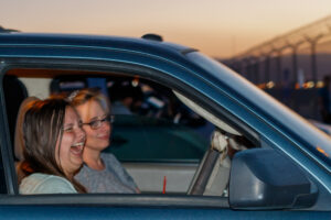 A woman and a girl sit in a dark blue car in a parking lot, laughing.