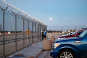 A man and a boy stand in front of a metal fence with a row of cars behind them.