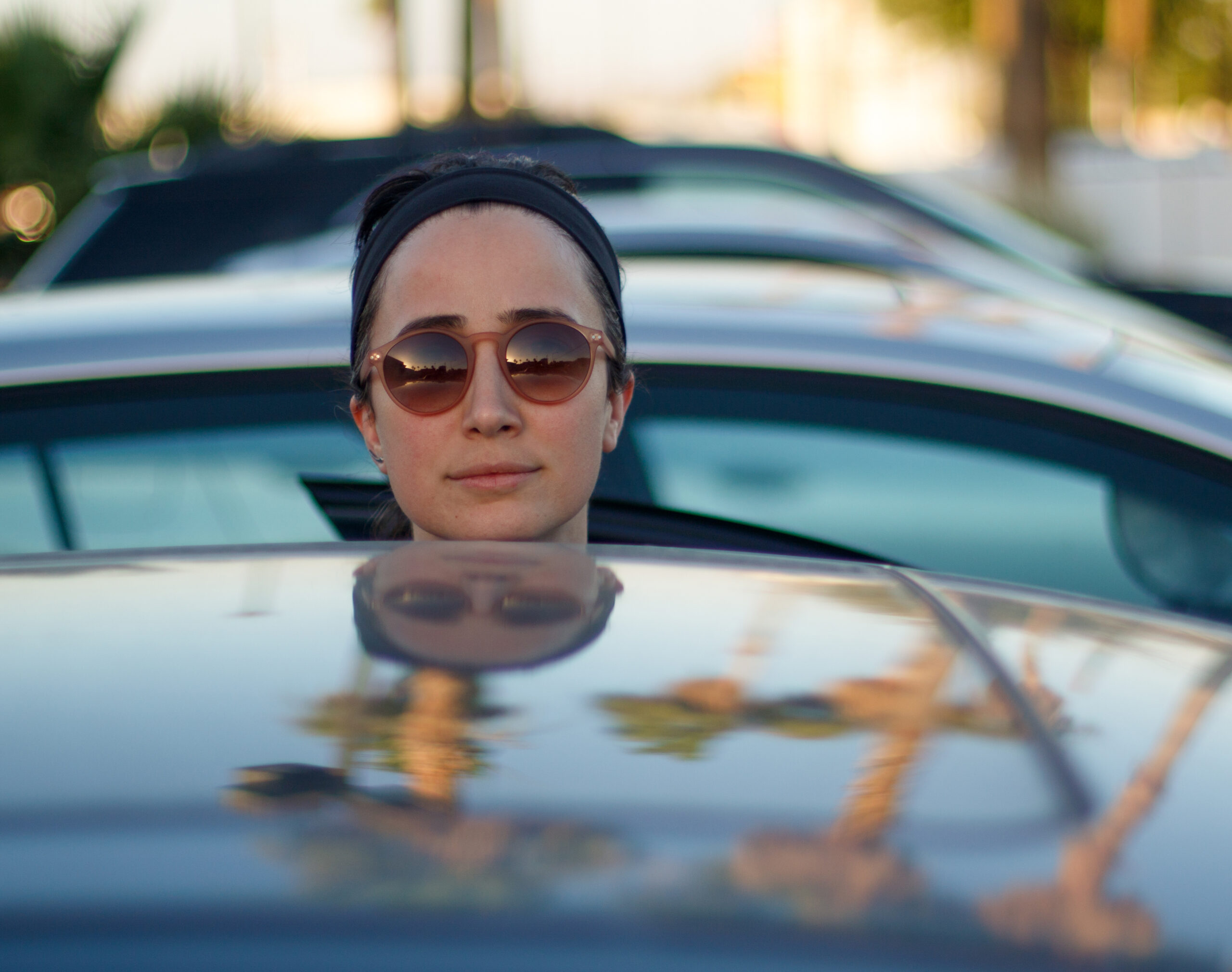 Woman wearing sunglasses sticking her head out from behind the roof of a car, which reflects palm trees in the sunlight.