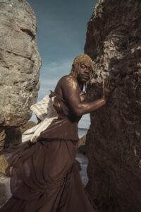jaamil olawale kosoko's headshot: a black person wearing a brown dress, standing between two rock formations.