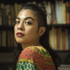 Ingrid Rojas Contreras's headshot: brown woman in an orange-green shirt against a background of books.