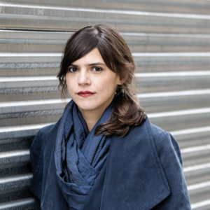 Valeria Luiselli's headshot: brown woman in a blue jacket against a metal grate. 