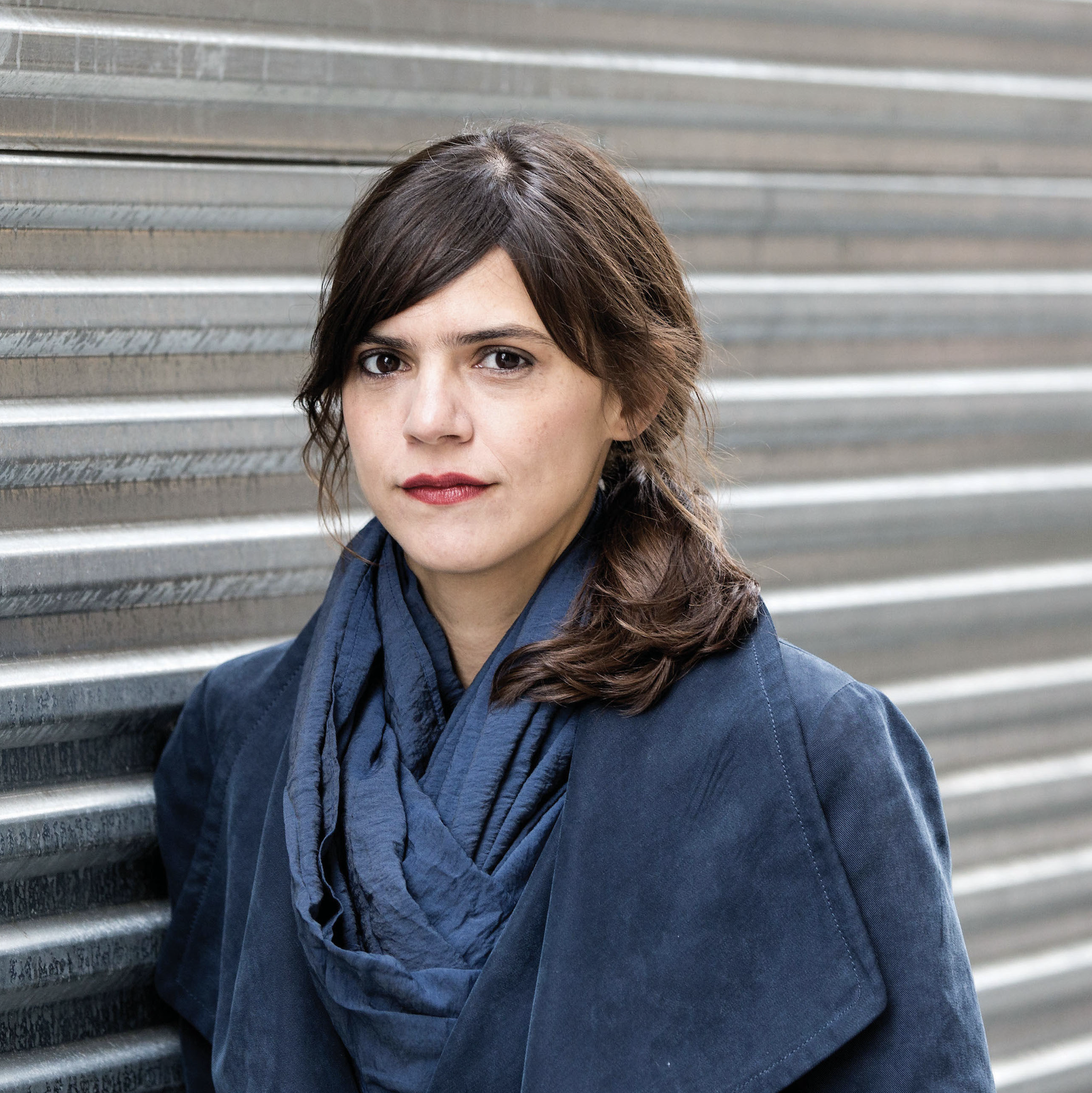 Valeria Luiselli's headshot: brown woman in a blue jacket against a metal grate.