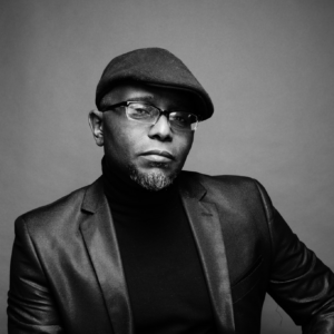 Tyehimba Jess's headshot: a black man wearing a suit and cap, black and white image.