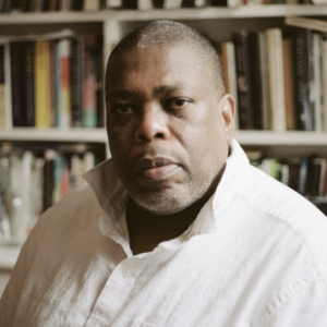 Hilton Als's headshot: black man in a white shirt against a background of books.