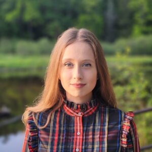 Sofia Belimova's headshot: white woman wearing plaid against a background of trees.