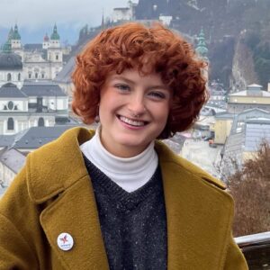 Image of Sam Spratford: White woman with red hair in a mustard jacket with a city in the background.