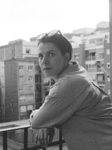 Sara Freeman's headshot: woman standing on a balcony against the background of apartment buildings
