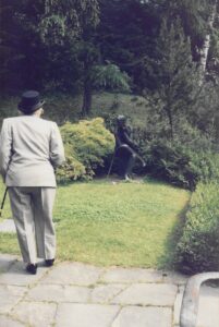 Elderly woman in a suite and top hat facing away from the viewer and towards a grave surrounded by bushes.