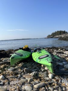 South Bristol, Maine. Two neon green Kayaks lay on shore atop rocks and pebbles, just before a body of water. Blue skies are above. Land is visible in the distance. 