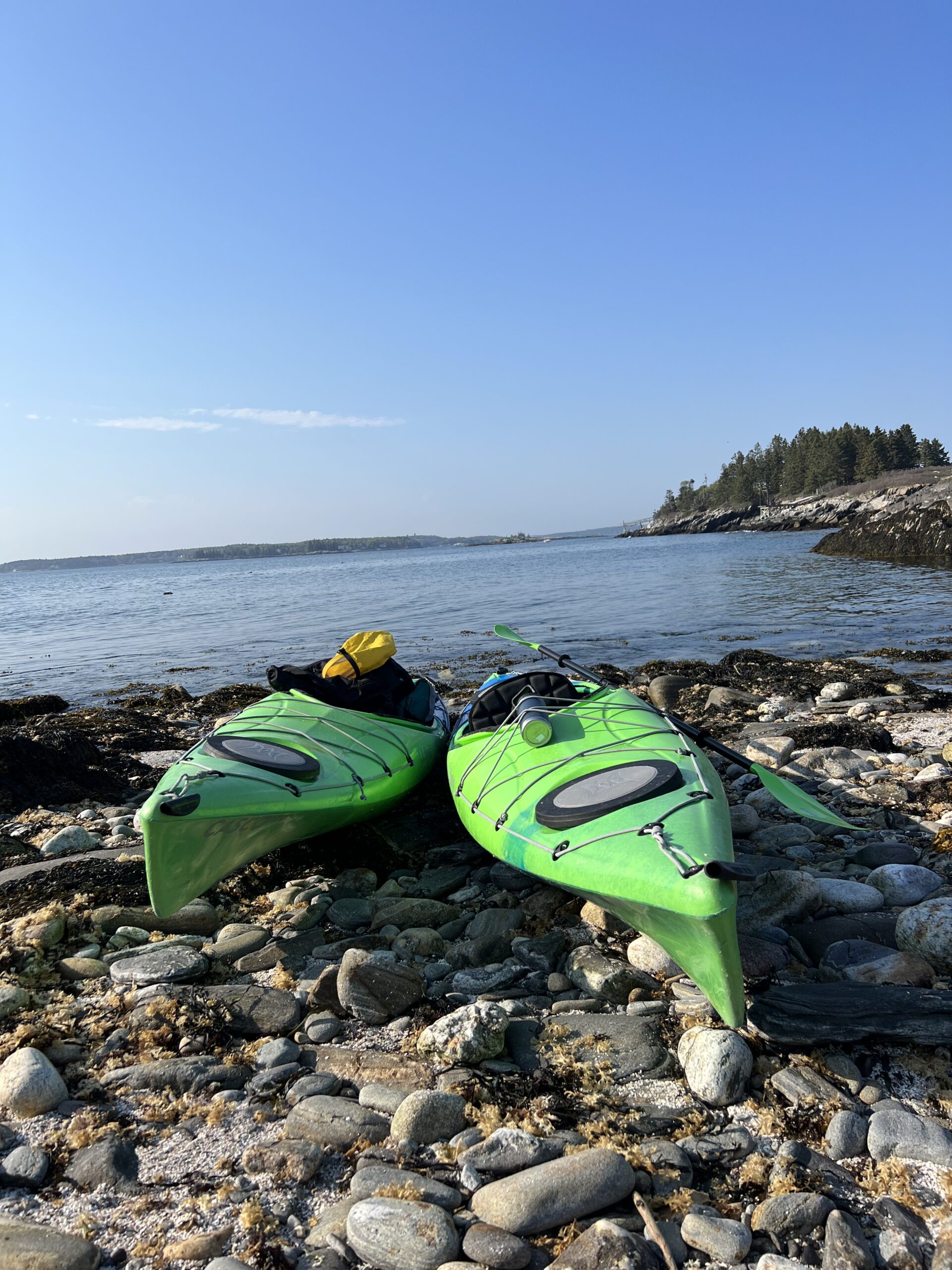 South Bristol, Maine. Two neon green Kayaks lay on shore atop rocks and pebbles, just before a body of water. Blue skies are above. Land is visible in the distance.