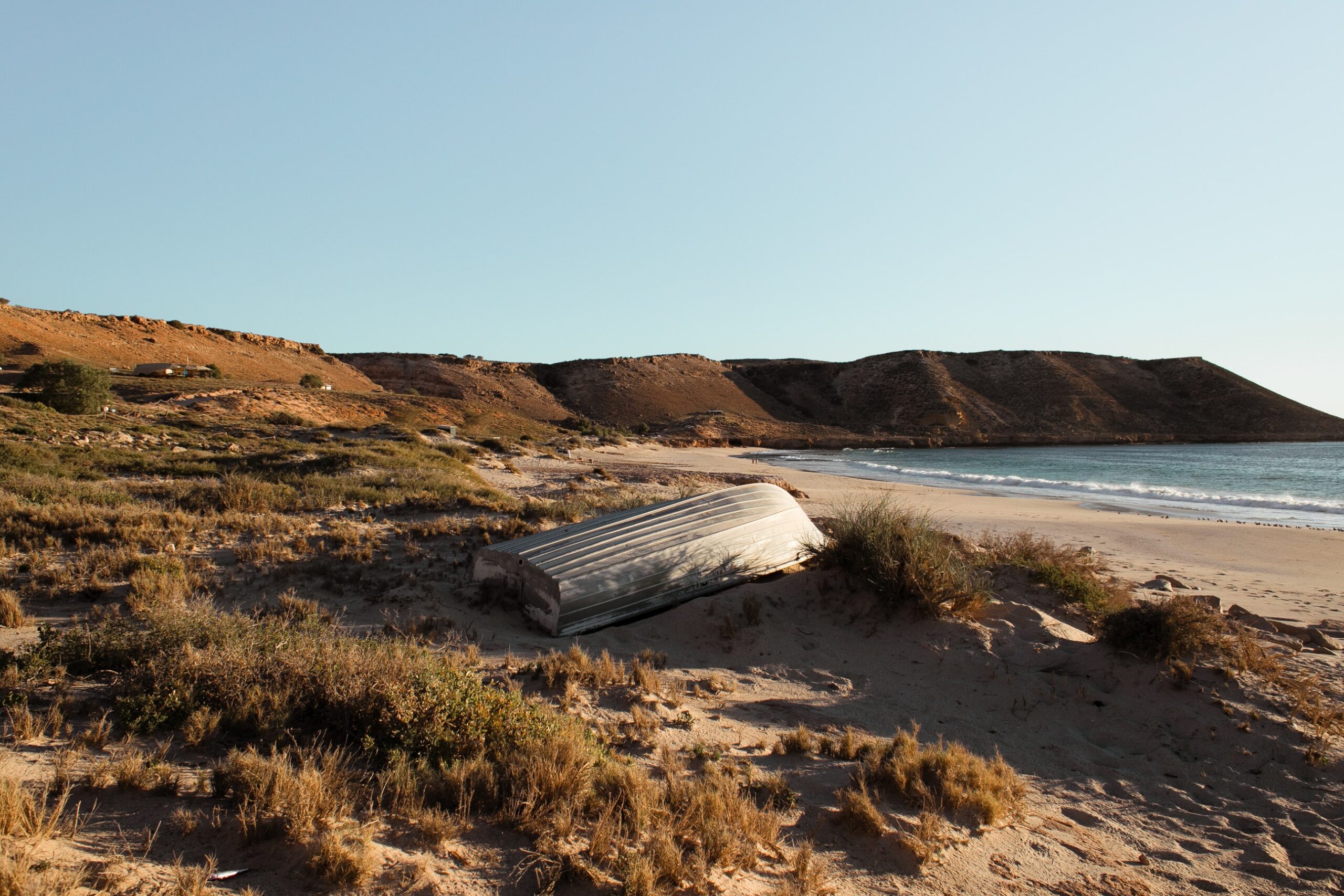 A flipped silver boat sits on a shore of dried grass and sand.