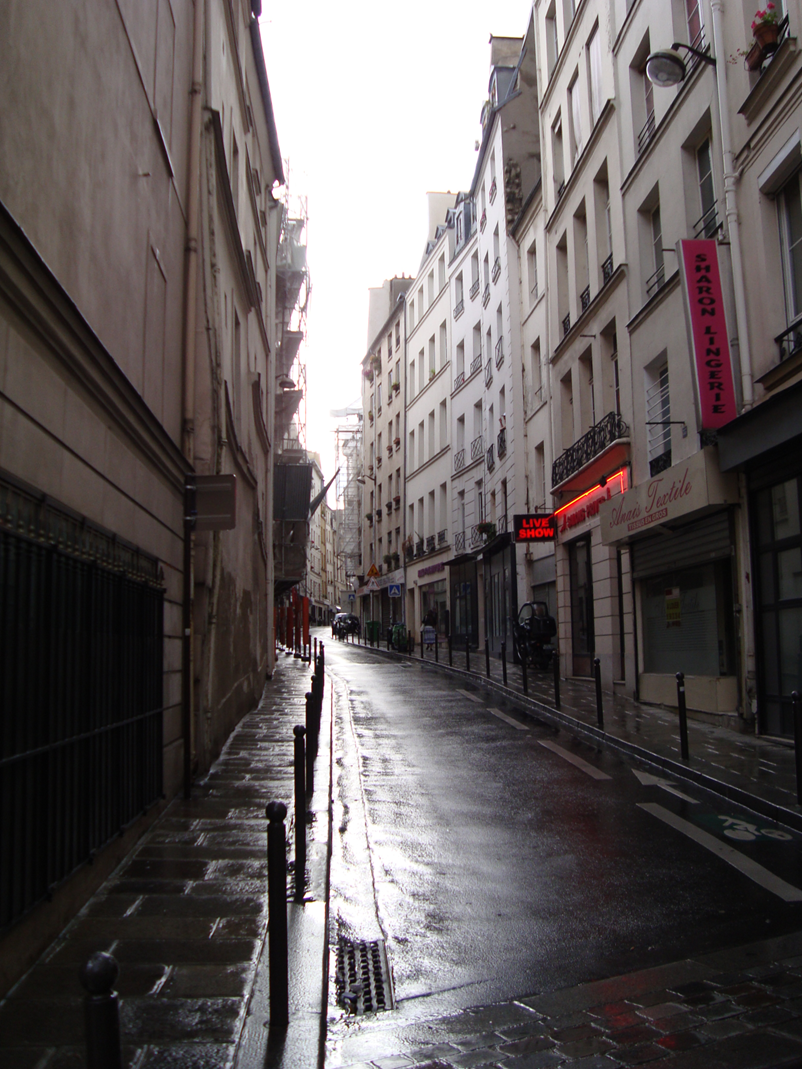 A narrow Paris street, sloping upward and slightly to the left, is slick with rain. Beige and white apartments frame the road, along with several red signs on the right advertising a "Live Show" and "Lingerie".