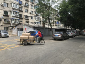 An apartment complex in Shenyang, Dongbei (China). A man rides a bike full of cardboard boxes in the foreground. Parked cars line the streets. 