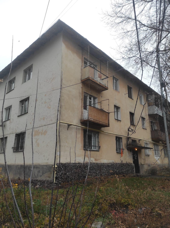 A rectangular, beige apartment building squats under an overcast sky. Dead branches and leaves crowd the foreground.