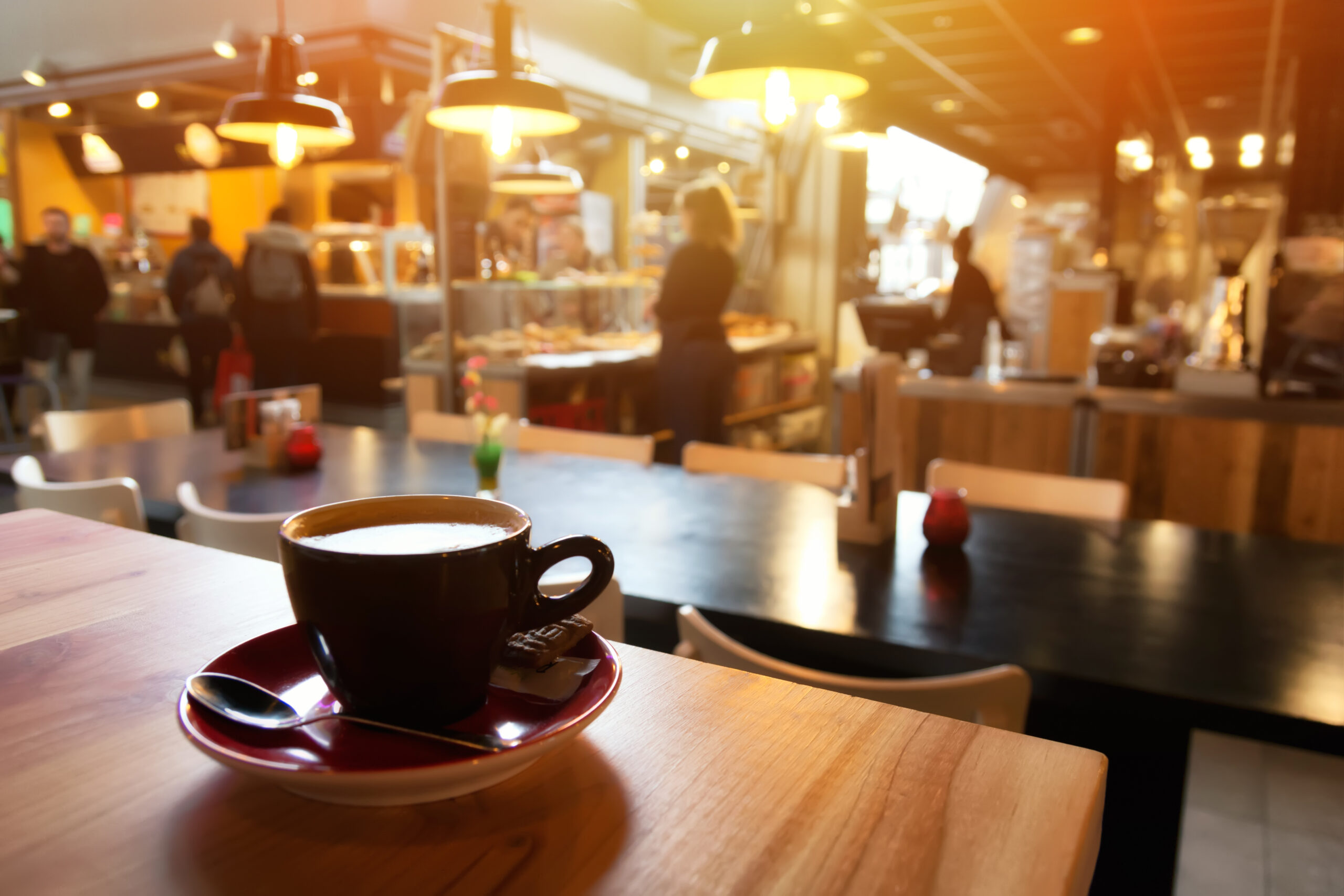 A coffee shop at sunset. The foreground is focused on a cappuccino; the background is blurred out of focus. People sit and talk.
