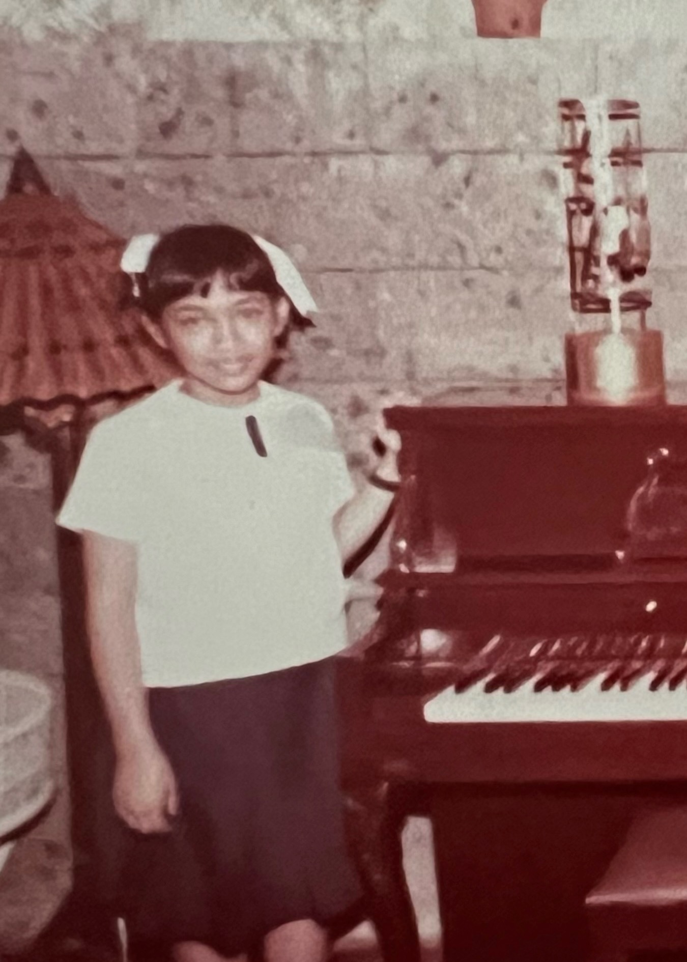 A sepia-tone image showing a young girl with a ribbon in her hair smiling next to a piano, one hand affectionately resting on the edge of the lid.