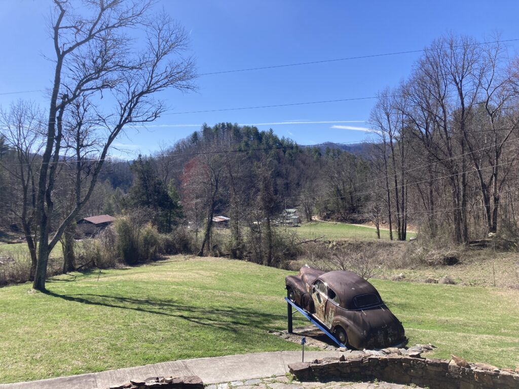 a broken down, rusty car  faces out toward a lightly forested, sunny, and hilly landscape.